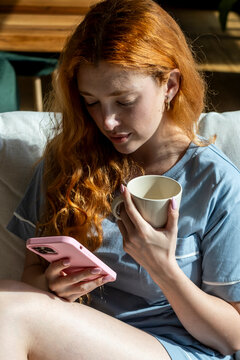 Redhead woman relaxing on a sofa in casual pajamas, holding a mug of coffee and checking her smartphone in warm morning sunlight, cozy at-home moment and quiet leisure