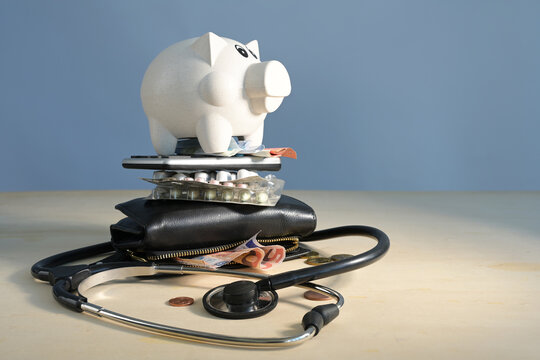 Healthcare costs symbolized by a stack of stethoscope, wallet, bills, pills, calculator and a piggy bank on top, light wooden table, blue background, home finance budget concept, copy space