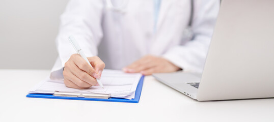 Fototapeta premium Professional doctor wearing lab coat and stethoscope using laptop computer and taking report notes on notebook at desk in hospital clinic office, medical technology and healthcare concept