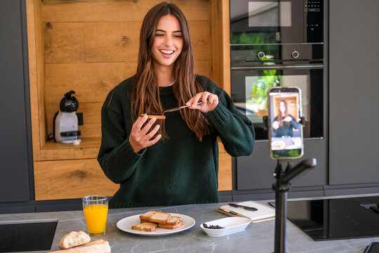 Young woman recording a breakfast vlog in a modern kitchen while smiling and spreading jam on toast, using a smartphone on a tripod to create food and lifestyle content