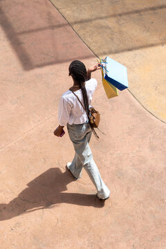 top view of afro woman with long dark braids, wearing a white lace blouse and jeans, standing with bags on a street or shopping center
