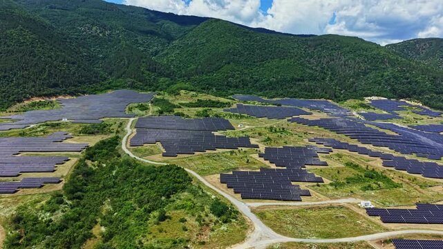 A large solar panel farm spans over hills in a valley with numerous solar panels aligned in rows. The sun shines on the panels as clouds drift by, showcasing clean energy production.