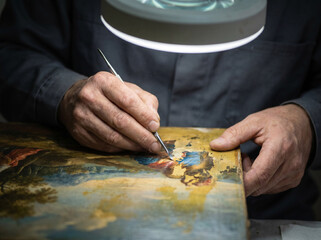 Fototapeta premium Art restorer's hands working on darkened old painting as original vivid colors emerge and brighten beneath specialized tool revealing centuries hidden beauty