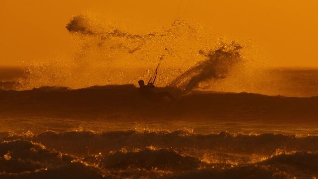 Man kiteboarding at sunset carves and snaps back on wave, telephoto slomo