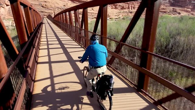 Aerial drone video follows a senior man cycling on the rustic Moab Canyon Bridge above the Colorado River in Moab, Utah