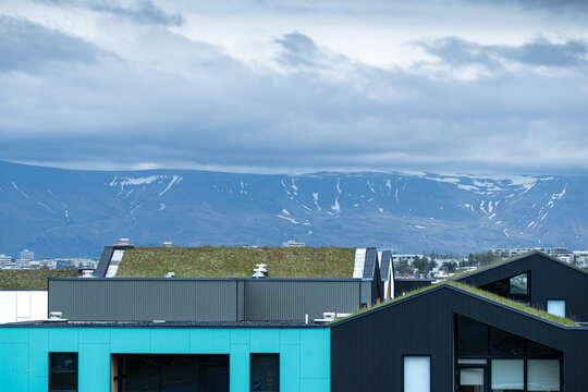 View of modern architecture with green sod roofs and colorful facades against a backdrop of snow-dusted mountains and a cloudy sky in Reykjavik, Reykjavikurborg, Iceland.