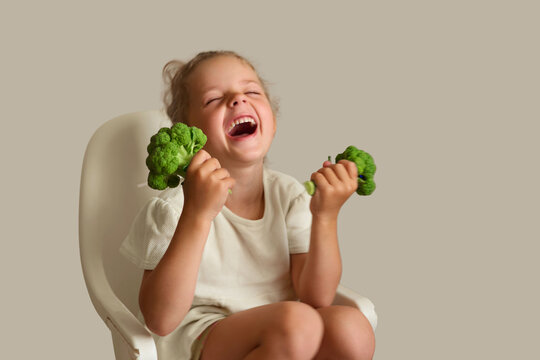 A young girl sits on a chair, holding two pieces of broccoli in her hands. She is laughing and shows joy while surrounded by a simple indoor setting