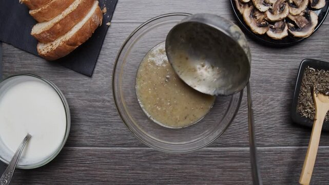 4K Video of chef pouring hot, delicious homemade mushroom cream soup with ladle into clear glass bowl on rustic wooden table with ingredients like sliced mushrooms, bread, sour cream, and spices.