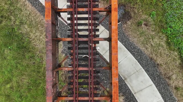 A bike pathway winding below an old rusty rail bridge spanning over a natural flowing creek and pedestrian bridge. Drone view
