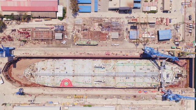 Massive tanker in a dry dock undergoing maintenance and repair, shown from a top-down aerial view with cranes and heavy machinery lining the shipyard and hull