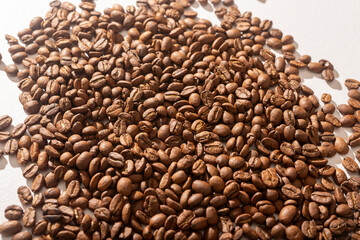 Obraz premium A pile of roasted coffee beans sits on a white background forming a natural mound shape. The heap of dark beans spills slightly against a clean light surface. Concept of coffee