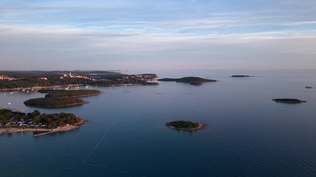 Pine-covered Adriatic islands scatter across calm blue sea off Vrsar coastline, with historic hilltop town and sailboat marina glowing in soft evening light. Croatia. Aerial drone approaching shot