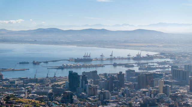 Aerial view of the bustling Cape Town harbor with container ships, cranes, and the urban skyline set against the backdrop of distant mountains in Cape Town, Western Cape, South Africa.