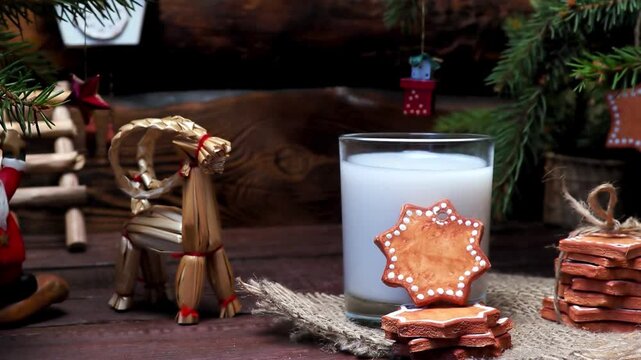 Christmas eve 4K video showing milk glass and star gingerbread cookies on burlap, rustic wooden table background with fir branches, traditional straw goat, santa claus toy figurine, retro cuckoo clock