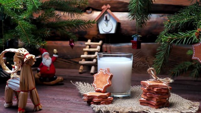 Christmas eve 4K video showing milk glass and star gingerbread cookies on burlap, rustic wooden table background with fir branches, traditional straw goat, santa claus toy figurine, retro cuckoo clock