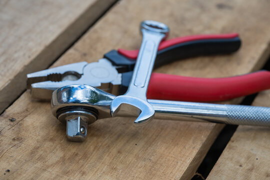 a pair of red-handled combination pliers, a chrome socket wrench, and an open-ended spanner lay gathered on a rough wooden board surface.