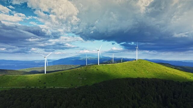 Wind turbines are placed on a green hill surrounded by trees. Dark clouds cover part of the sky as sunlight shines through. Mountains are visible in the background.