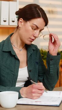 Young woman on home table writes notes with pen removes glasses drinks coffee thinks during study break in living room. Adult lady on home table puts glasses back and continues writing for work task.