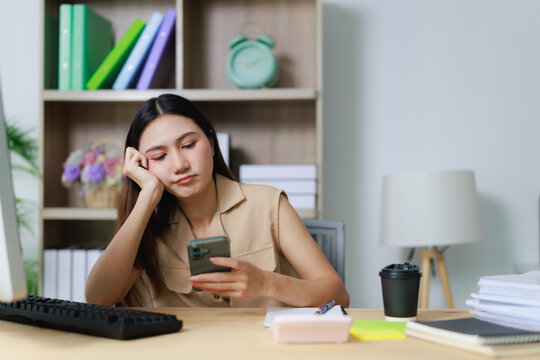 Young Asian woman feeling disengaged during her workday, using her mobile phone instead of focusing on tasks, showing signs of weariness and lack of motivation