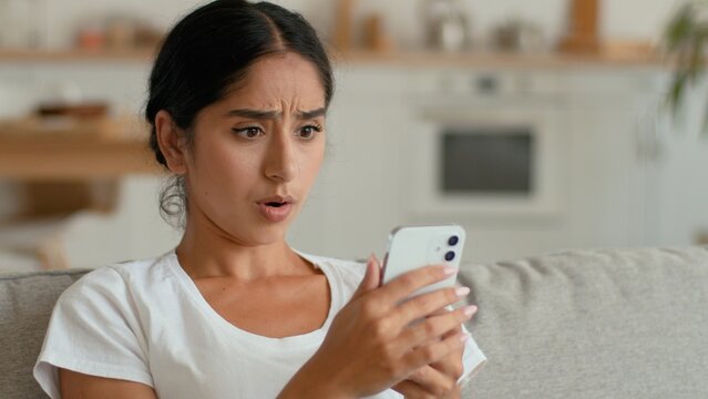 Shocked Indian Arabian woman at home using mobile phone gadget addicted social media obsessed webcam ethnic female girl taking selfie photo vlogger influencer dissatisfied surprised upset indoors