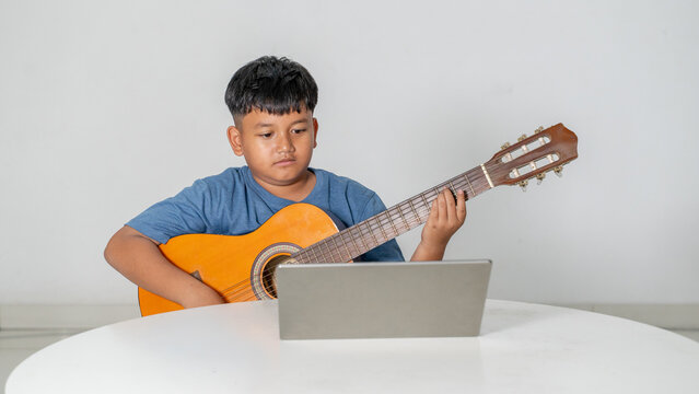 Focused Young Asian Boy Learning to Play Acoustic Guitar with Laptop Online Lesson