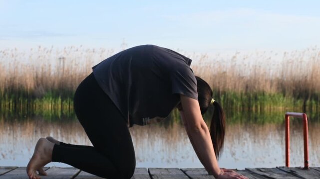 Woman performs tabletop yoga pose on wooden pier by calm lake in natural setting. Practitioner focuses on stretching, balance and mindful wellness. Woman expresses flexibility and inner calm.