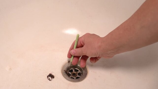 Hand pulls hair clog out of drain with metal tool. Dirty bathtub requires regular plumbing maintenance. Person solves hygiene issue by clearing dirty pipe hole. Domestic routine for cleaner home.