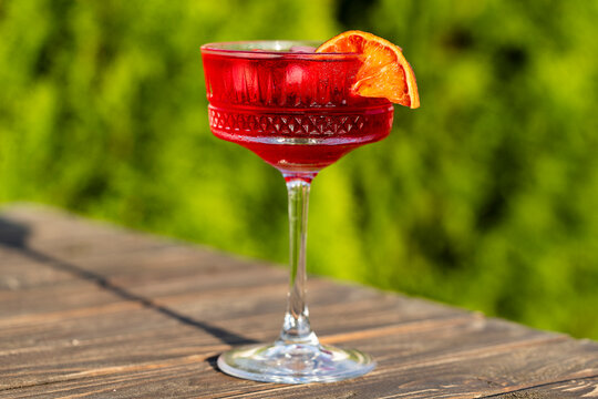Blood orange margarita cocktail in coupe glass on wooden table. Refreshing red negroni or campari cocktail with ice cubes and orange garnish in a glass on a sunny day