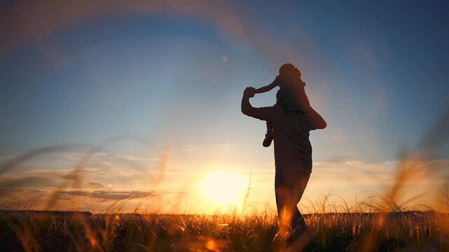 A man holds a child on his shoulders during sunset. The man and child are silhouetted against a colorful sky and tall grass, enjoying the moment together.