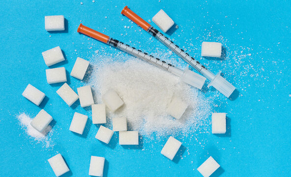 Two insulin syringes and scattered white sugar on a blue background, representing the problem of diabetes and glucose dependence.
