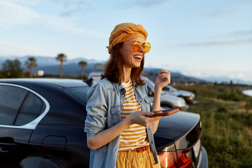 Fototapeta premium Smiling woman in striped shirt and denim jacket stands beside a car in sunny outdoor setting, capturing a lifestyle moment with relaxed mood and warm light.