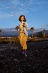 Fototapeta premium A young woman with curly hair walks along a rocky beach at golden hour, wearing a mustard dress and light jacket, smiling softly as palm trees frame the scene.