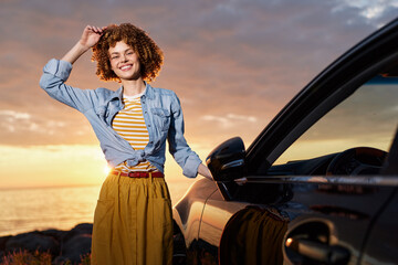 Fototapeta premium Joyful woman outdoors by a car at sunset, wearing a denim jacket over a striped shirt and a yellow skirt, smiling with wind in her hair, relaxed pose, warm tones