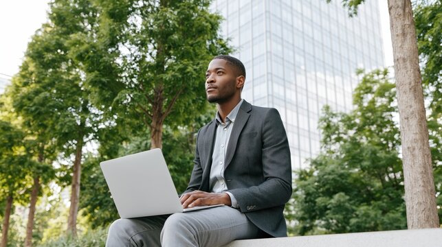 Young Black man in blazer working on laptop outdoors near modern building