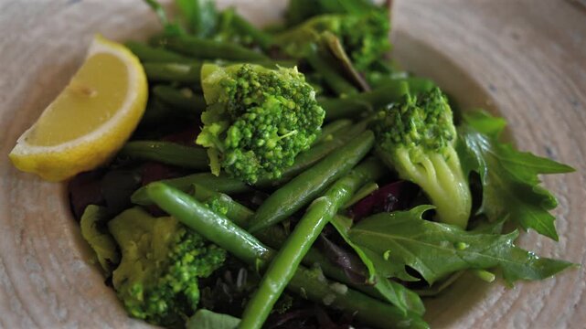 Green vegetable salad with fresh broccoli string beans and lemon wedge on ceramic plate in restaurant.