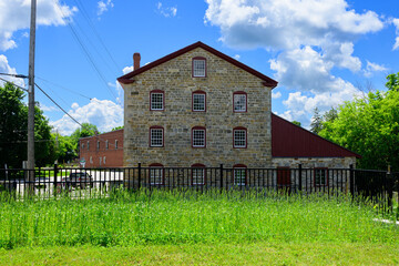 Fototapeta premium Historic stone mill behind wheat field | The historic Old Stone Mill stands behind a field of tall green grass and plot of heritage wheat under a blue sky with clouds in Delta, Ontario.