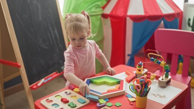 Little blonde girl places wooden puzzle pieces with her hand by a chalkboard and play tent inside a building; curiosity focus discovery.