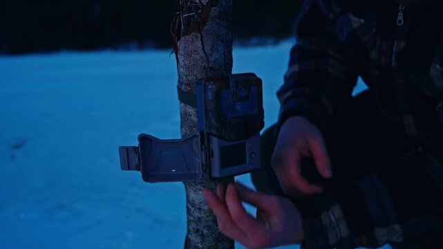 A Person Attaches a Trail Camera to a Tree Trunk in a Snowy Outdoor Environment During Low Light Conditions - Close-Up Shot