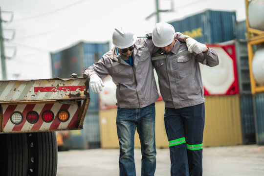 Senior worker shows signs of heat exhaustion while younger coworker provides support near truck trailer. Logistics operations workplace health remains important in hot outdoor conditions.