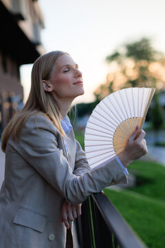 Young businesswoman cools down with a fan during heatwave while standing on balcony in evening light
