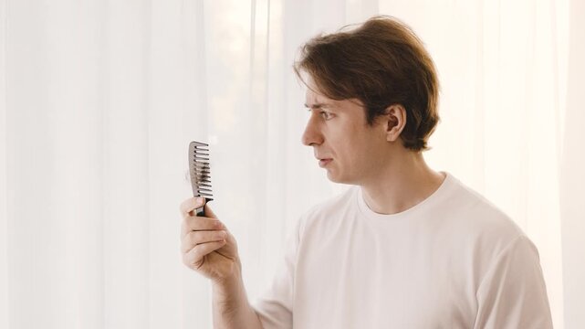 Worried man examines a comb with fallen hair. This male grooming concept shows alopecia, testosterone changes, and stress-related hair loss. High quality 4k footage