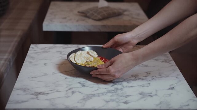 A pair of hands carefully placing a black bowl containing a healthy breakfast dish onto an elegant white marble-patterned table. 