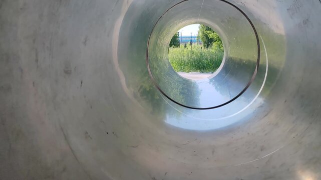 metal pipe creates circular tunnel view to green meadow. this pipe provides unique cylindrical perspective of outdoor landscape. metal pipe frames natural scenery