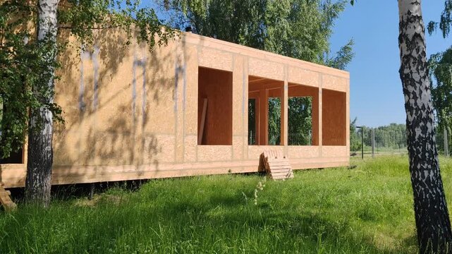 wooden frame house is under construction with OSB panel walls. window and door openings are cut in the structure. this house is built on a wooden foundation surrounded by green grass