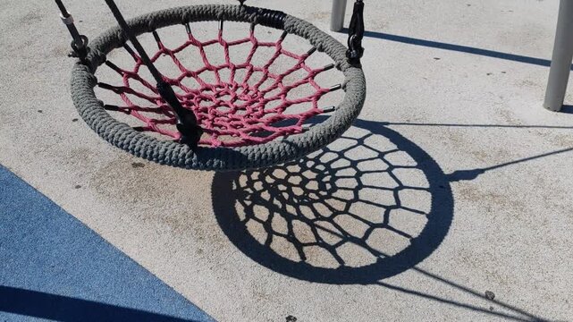 round spider web swing hangs above rubberized playground surface. bright sunlight casts distinct net shadow on the ground. Empty circular rope nest swing hanging on playground, casting sharp shadow
