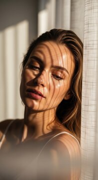 Close-up of a young woman with closed eyes and stripes of shadow from window curtains casting across her face during golden hour.