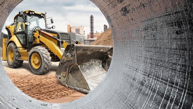 A backhoe loader is working at a construction site, moving dirt and preparing the ground for a new building in an urban area with factories nearby