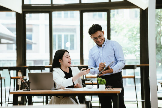 Smiling business colleagues collaborating at a modern office, reviewing work on a laptop while sharing ideas over coffee, highlighting teamwork, productivity, and professional communication.