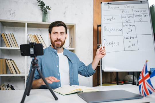 Bearded male tutor sitting at table next to whiteboard and recording a video