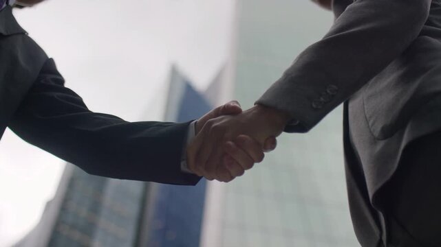 Close-up of business professionals shaking hands outdoors against a background of modern corporate skyscrapers, successful deal or partnership concept
